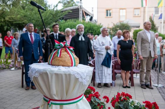 Kovács Péter polgármester Ács Anikó és Szász József alpolgármesterek, valamint a kerületi egyházi képviselők körében hallgatja a Himnuszt a Kertvárosi Olimpikonok Parkjában, előtérben a trikolórral átkötött újkenyér